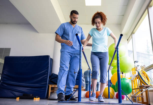 Male nurse assisting a lady walk. The lady is using two horizontal bars to hold herself up.